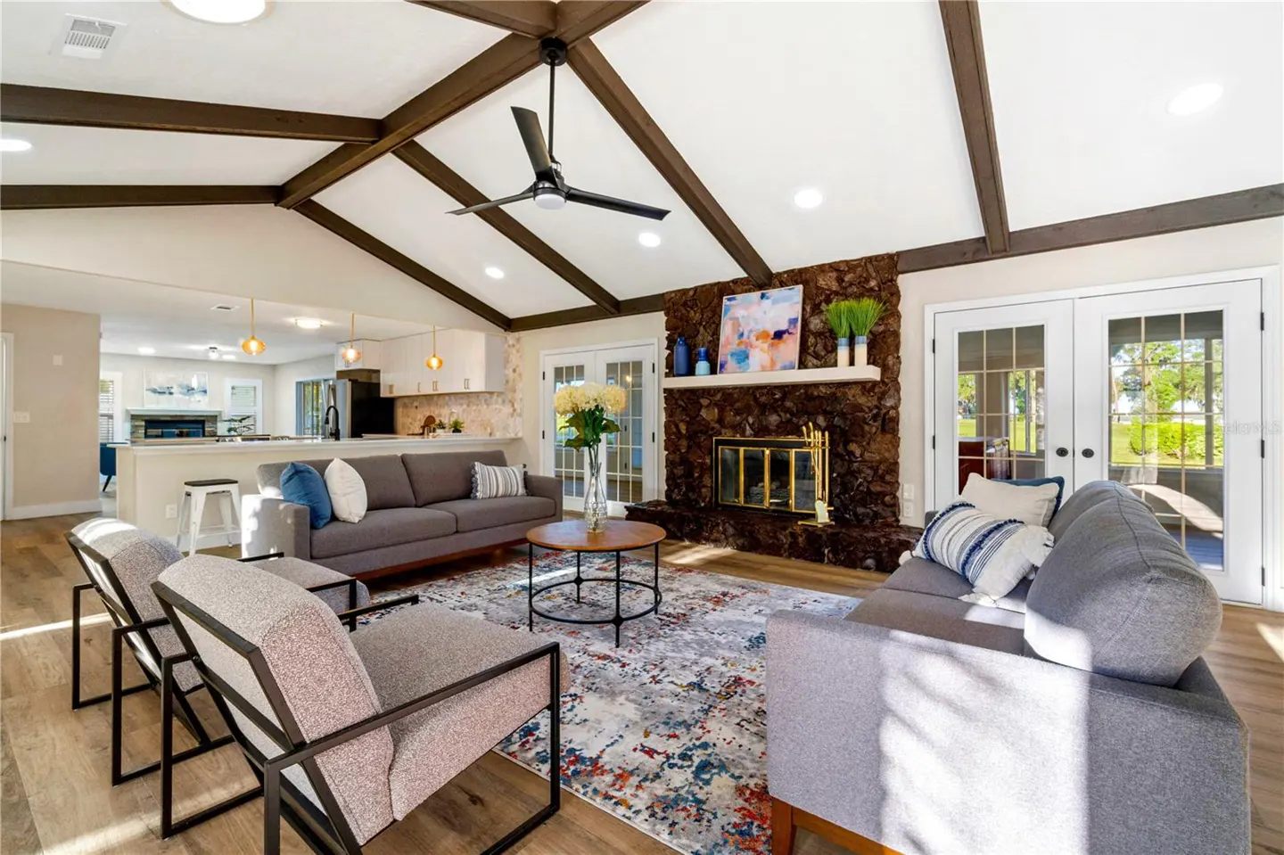 Bright living room with vaulted ceiling, wood beams, stone fireplace, gray sofas, and colorful rug.