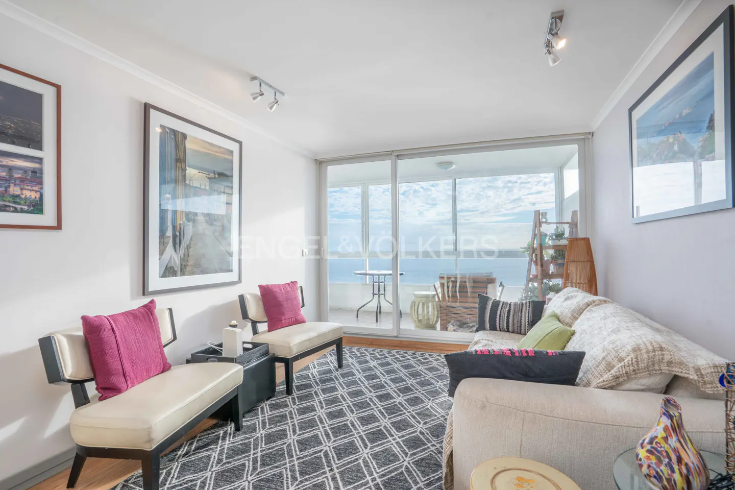 Bright living room with white walls, patterned rug, two cream chairs with pink pillows, and a sofa overlooking the ocean.