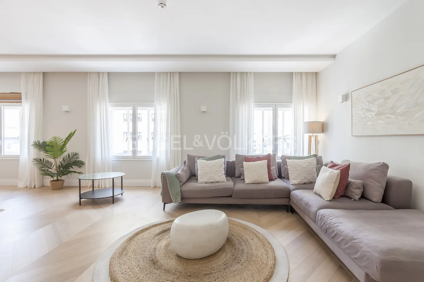 Bright living room with a gray sectional sofa, decorative pillows, a round jute rug, and a white ottoman. Windows with sheer curtains let in natural light.