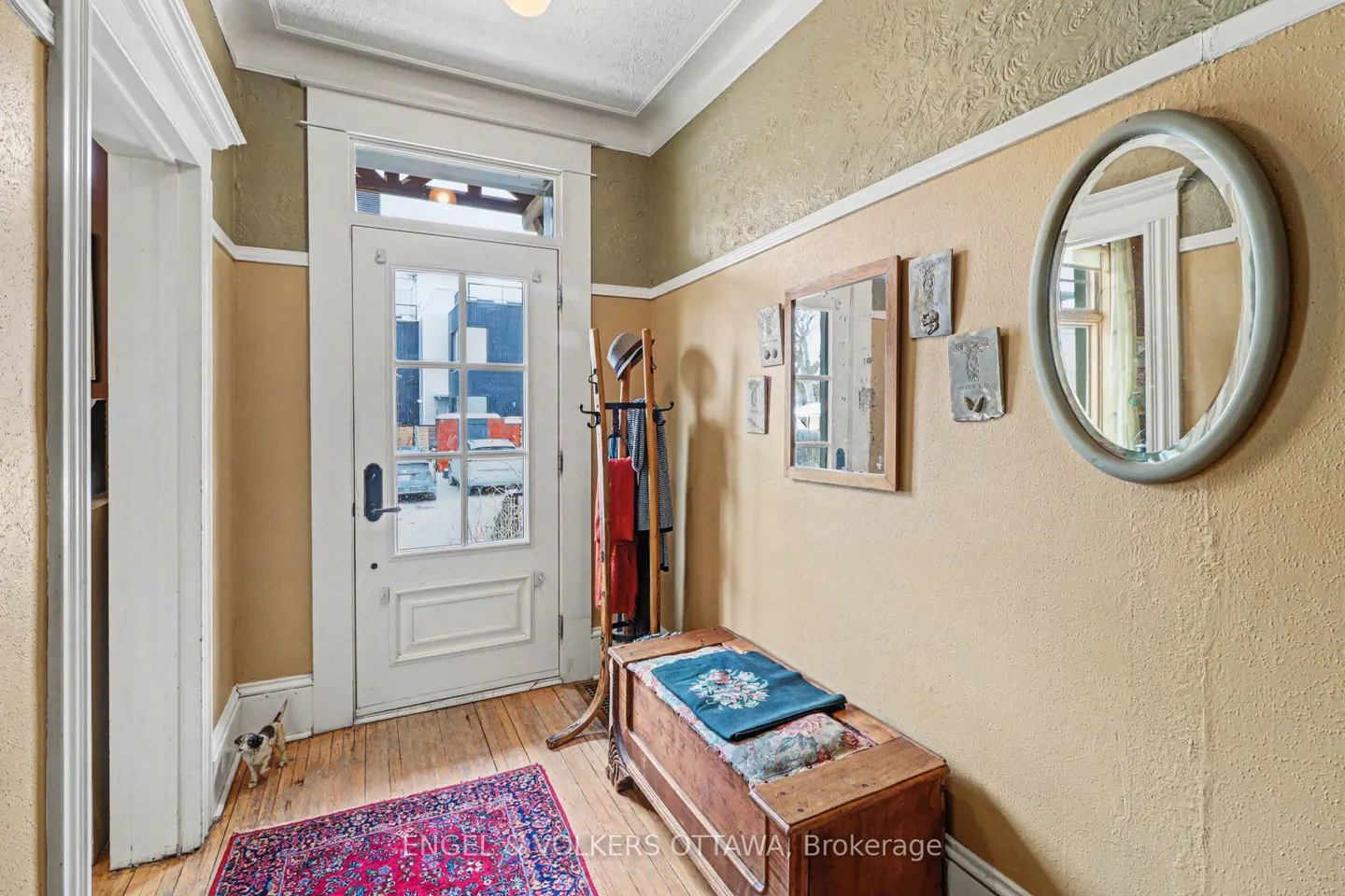 Foyer with a white door, wooden floors, and tan walls. A wooden chest sits near a coat rack. Mirrors and art hang on the wall.