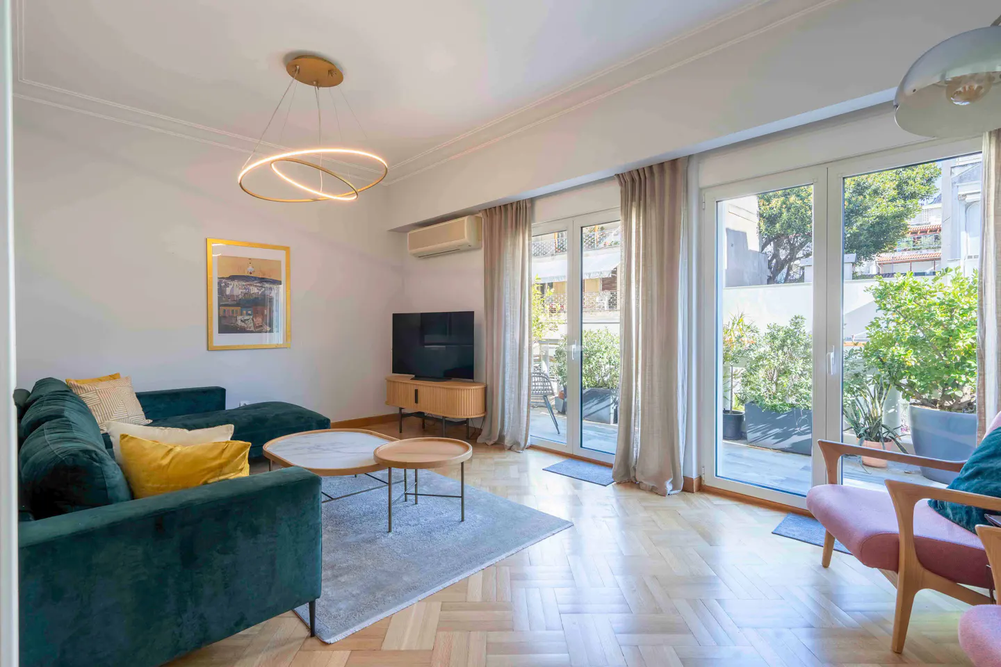 Bright living room with a green velvet couch, yellow pillows, and a round coffee table on a gray rug. French doors lead to a patio with plants.