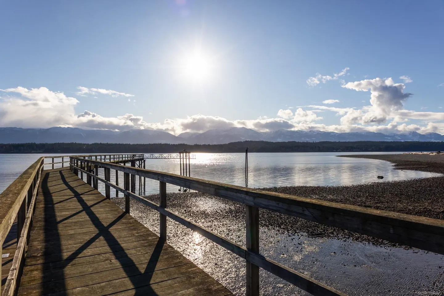 Wooden pier stretches into calm water under a bright sun and blue sky with clouds. Mountains are visible in the distance.
