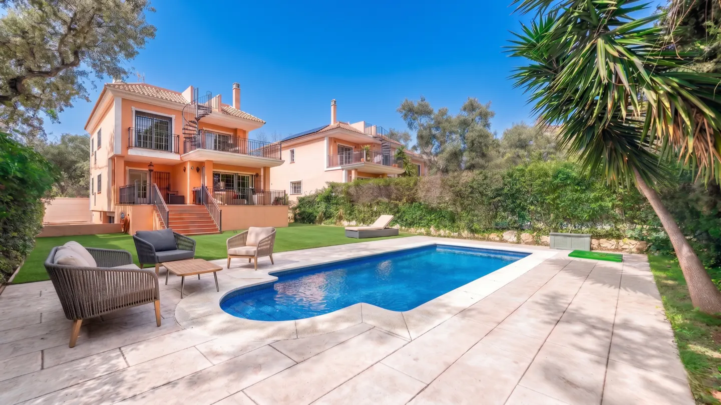 Backyard view of a two-story peach house with a blue swimming pool, patio furniture, and green landscaping under a clear blue sky.