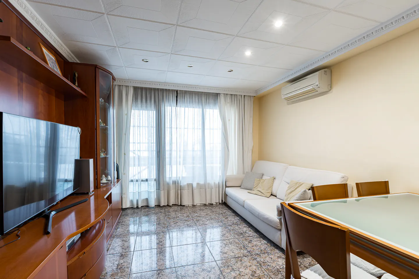 Living room with a white sofa, a wooden TV stand, and a dining table near a window with sheer curtains.
