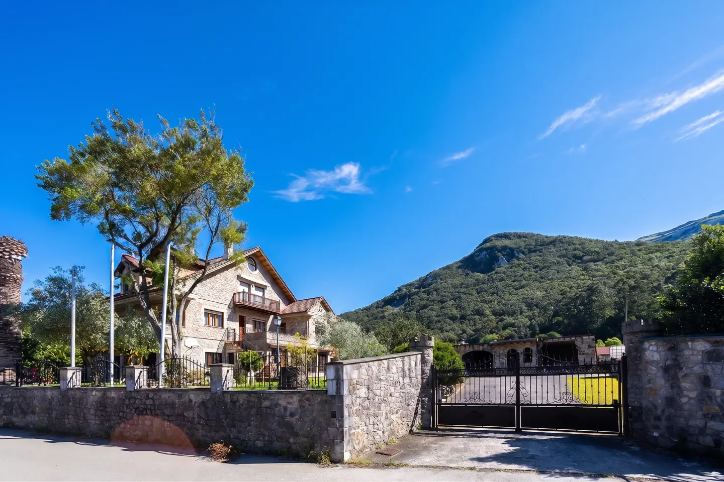 Stone house with a balcony and a tree, behind a stone wall and a black metal gate, with a mountain in the background.