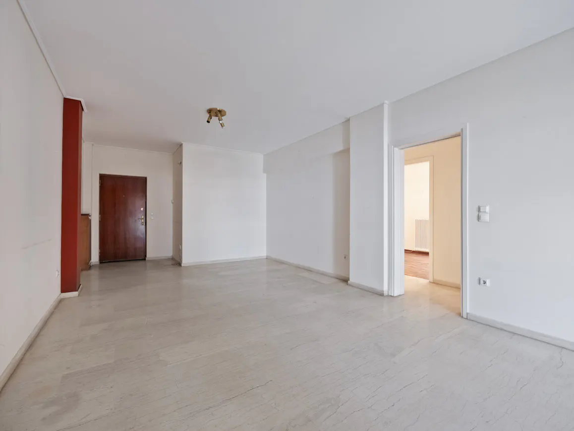Empty room with marble floor, white walls, and a dark wood door. A doorway leads to another room with a radiator.