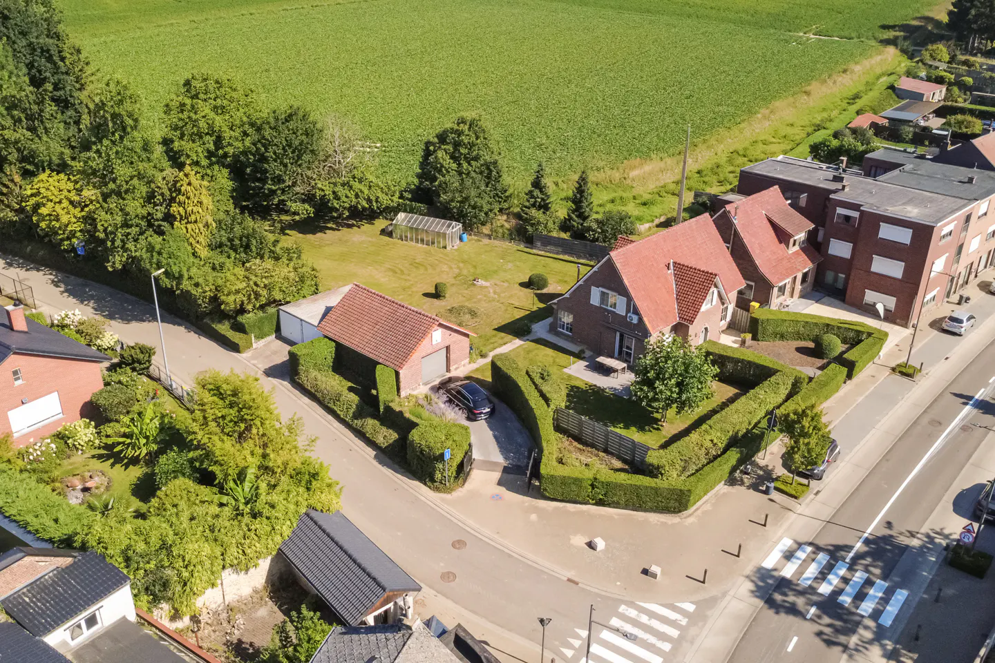 Aerial view of a brick house with a red tile roof, surrounded by green hedges and trees, next to a green field.