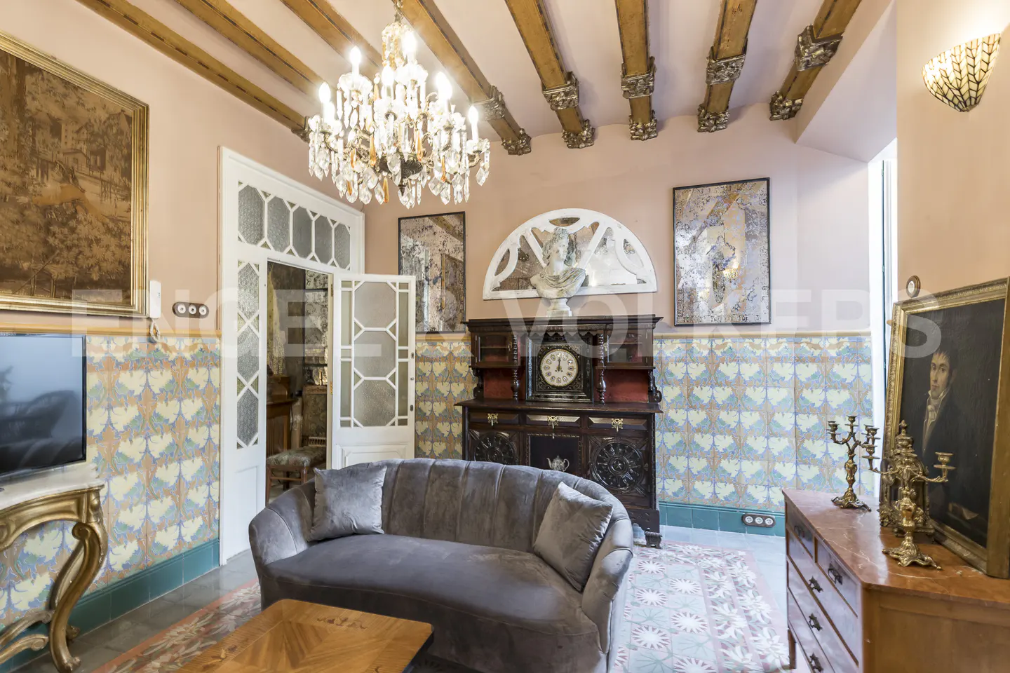 Ornate living room with pink walls, wooden beams, and a crystal chandelier. A gray sofa sits in front of a dark wood cabinet with a clock. Paintings adorn the walls.
