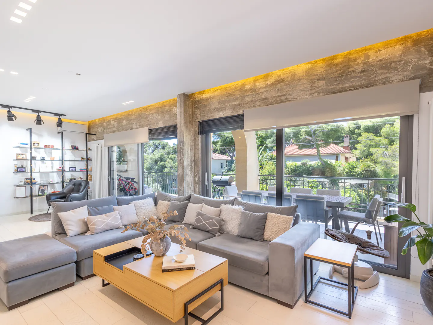 Modern living room with a large grey sectional sofa, wooden coffee table, and floor-to-ceiling windows overlooking a balcony.