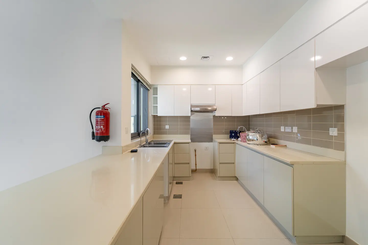 Bright kitchen with white cabinets, beige countertops, and a stainless steel range hood. A red fire extinguisher is mounted on the wall.