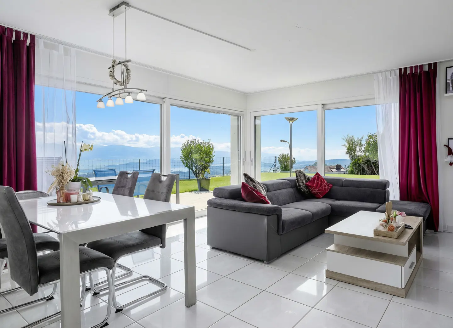 Bright living room with a white table, gray chairs, and a gray sofa. Large windows offer a scenic view of a lake and mountains.