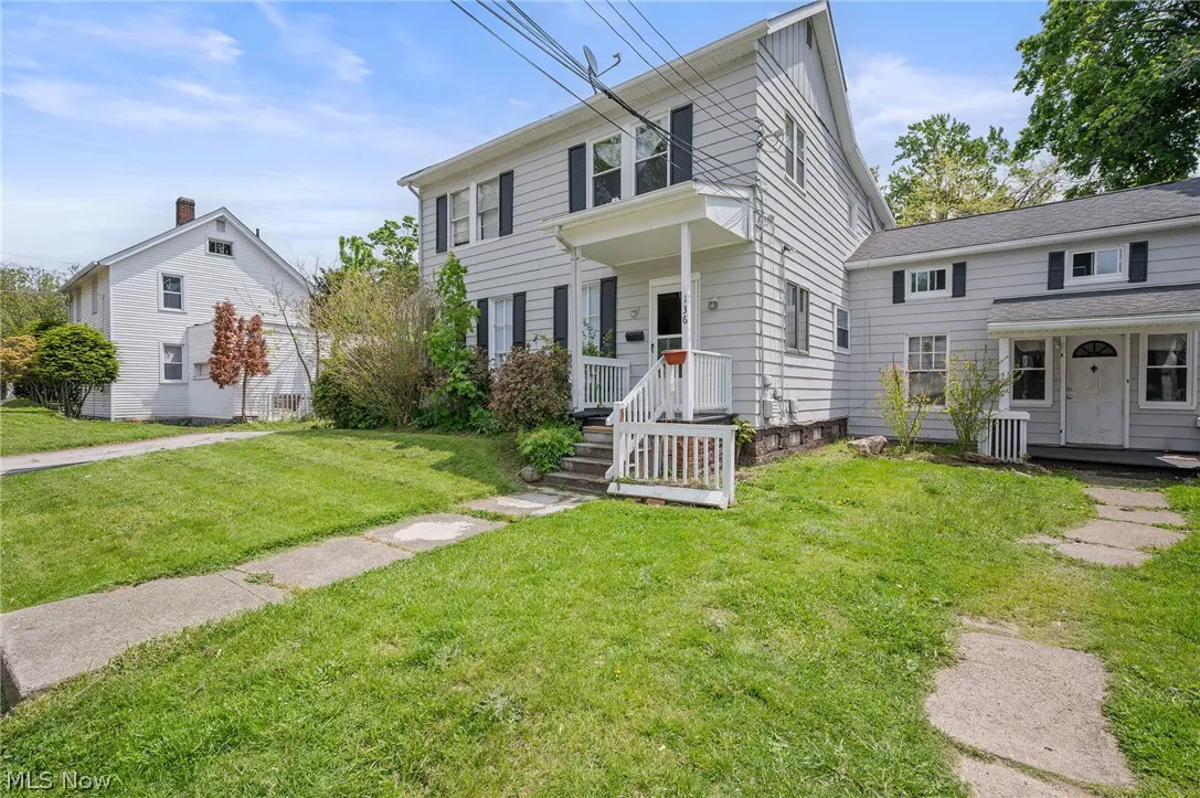 Exterior view of a two-story, light gray house with black shutters and a white porch. Green lawn and blue sky.