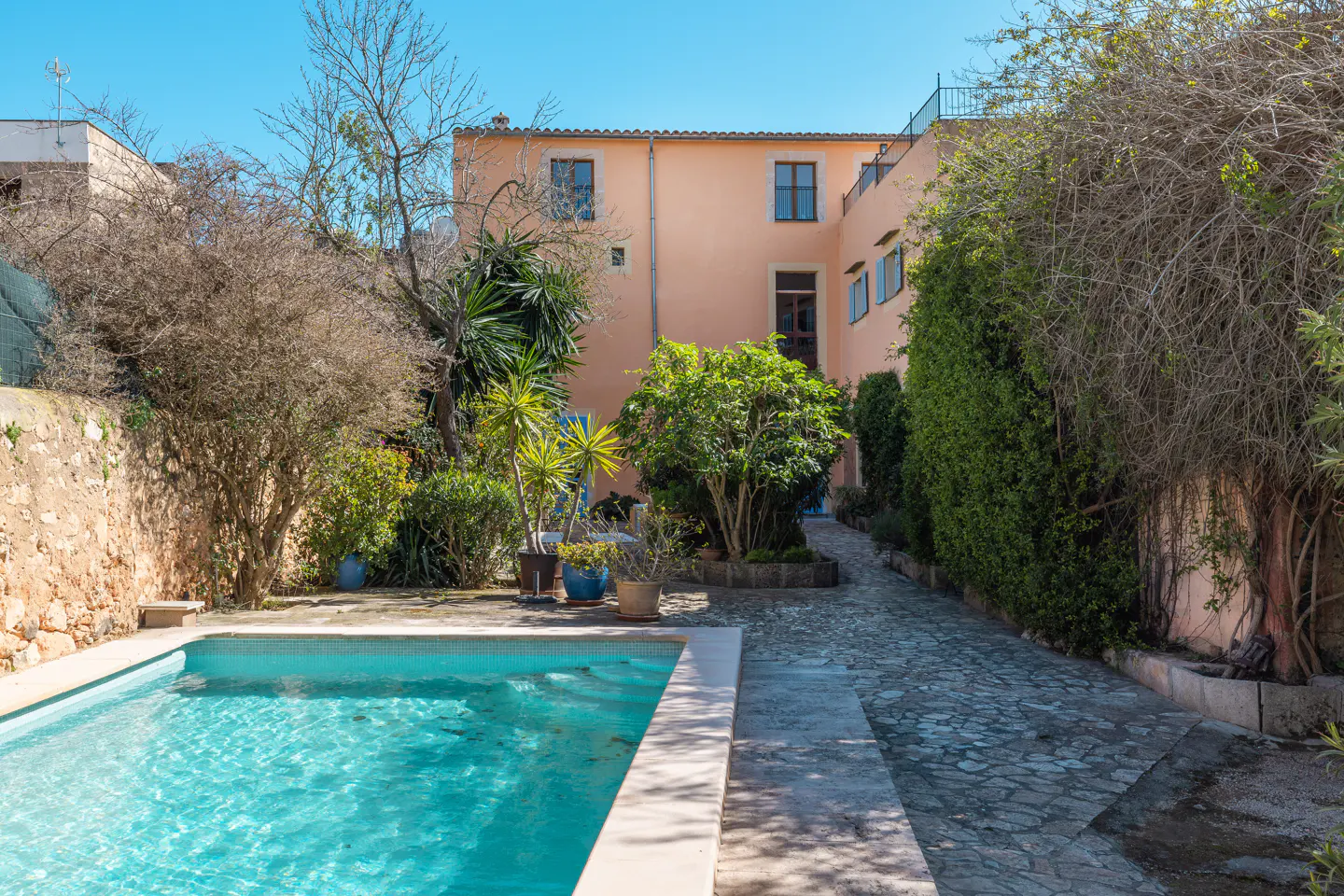 A peach-colored building overlooks a pool with turquoise water, surrounded by lush greenery and a stone path.