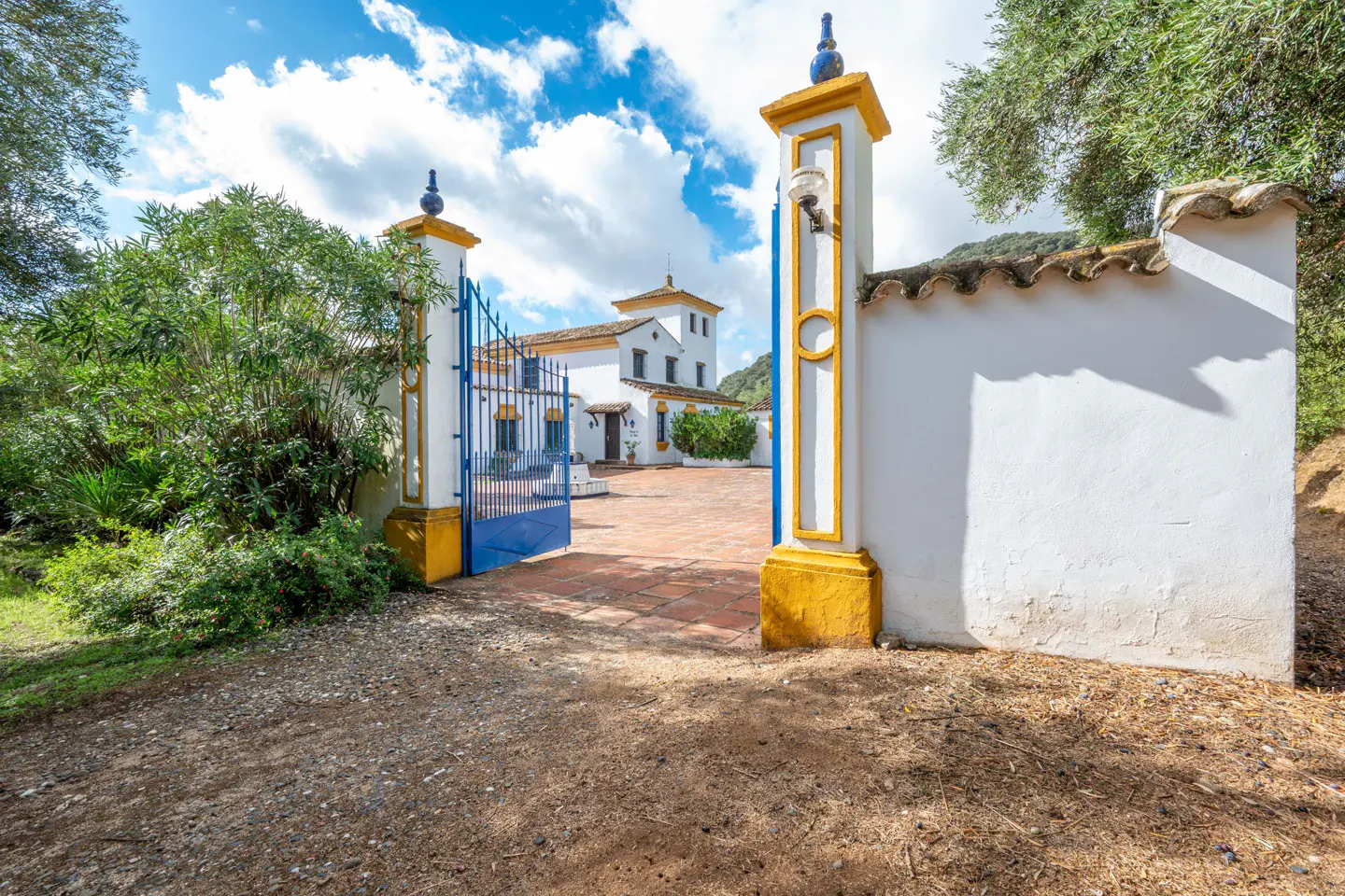 View through open blue gates to a white house with yellow trim and a red brick courtyard.