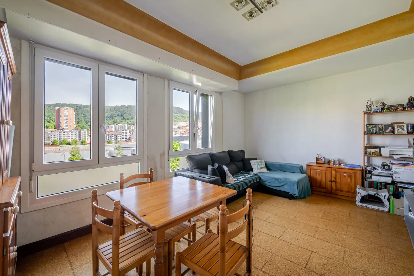 Bright living room with a wood table, chairs, and a gray sofa with a blue blanket. Large windows overlook a city.