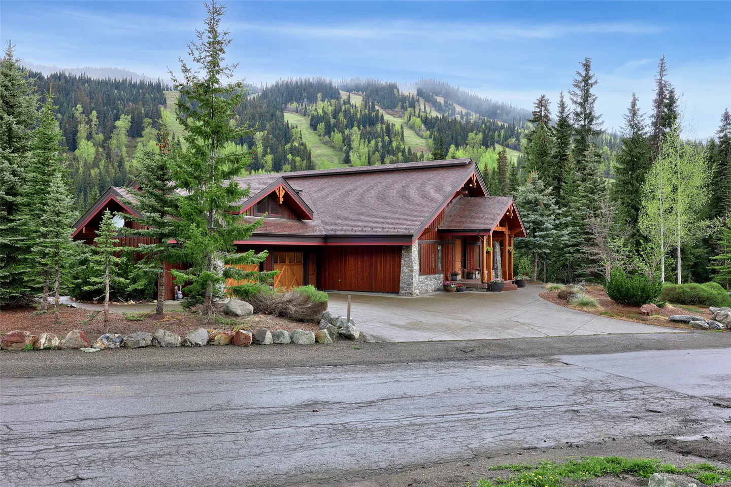 A wood and stone house with a brown roof sits in front of a mountain covered in green trees.