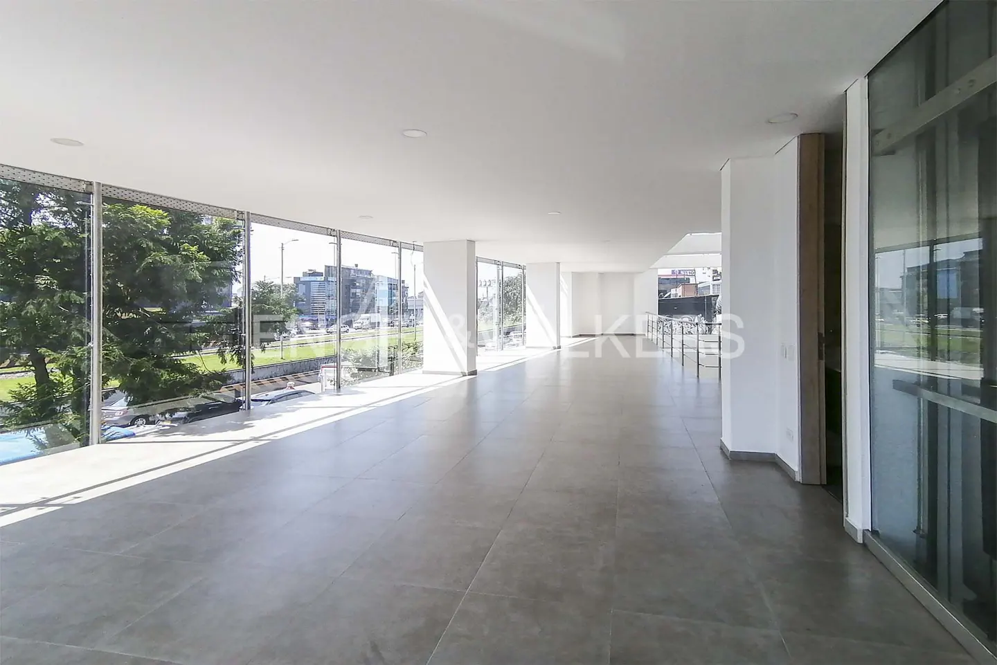 Bright, empty room with gray tile floor and floor-to-ceiling windows showing trees and city buildings.