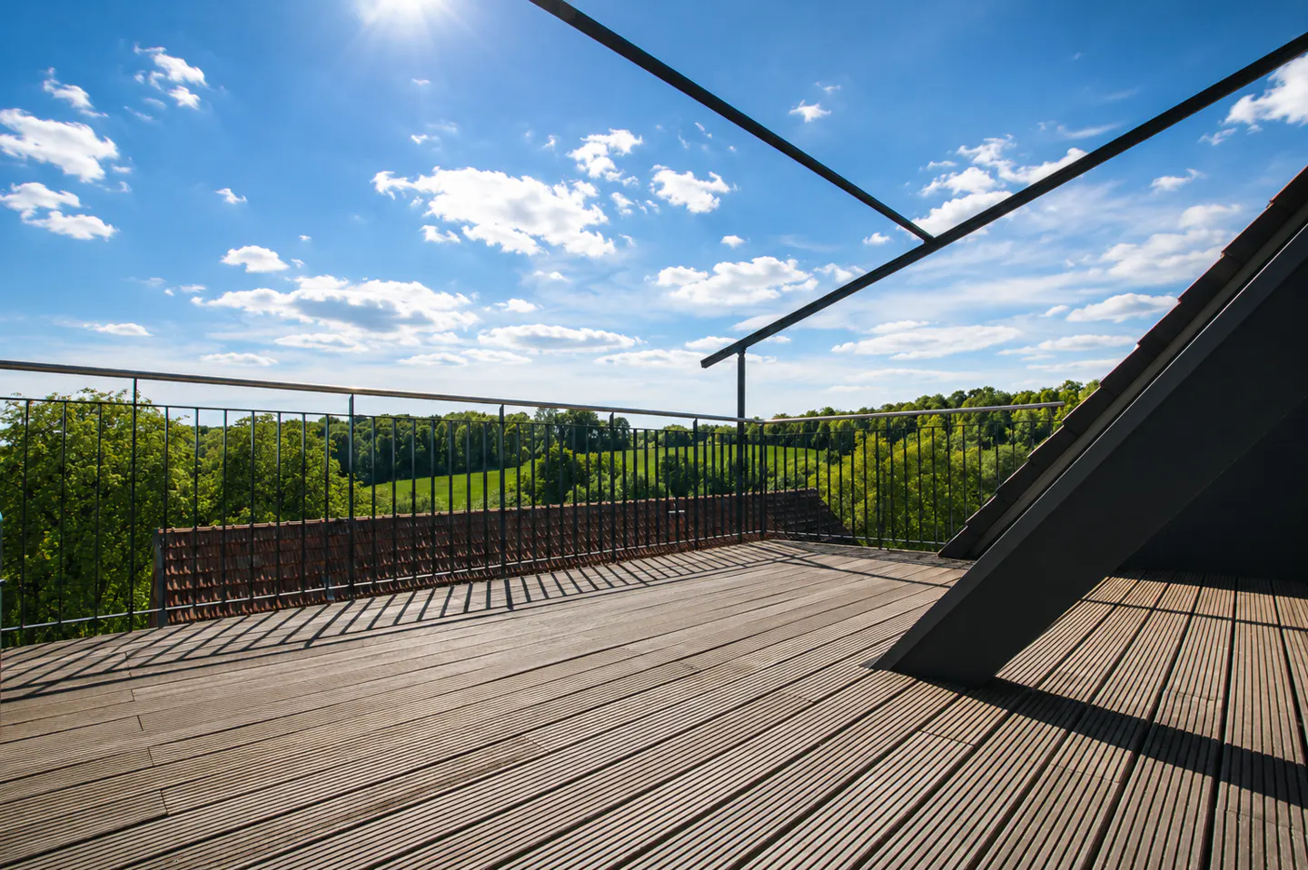 Wooden deck with black metal railing overlooks green trees under a blue sky with white clouds and sun.