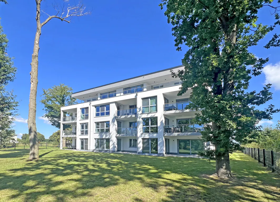 Modern white apartment building with balconies, set on a green lawn under a blue sky, framed by trees.