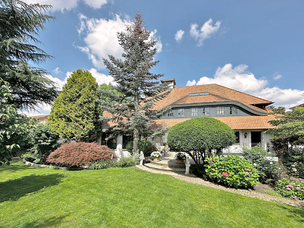 Exterior view of a large white house with a red tile roof, surrounded by lush green landscaping and trees under a blue sky.
