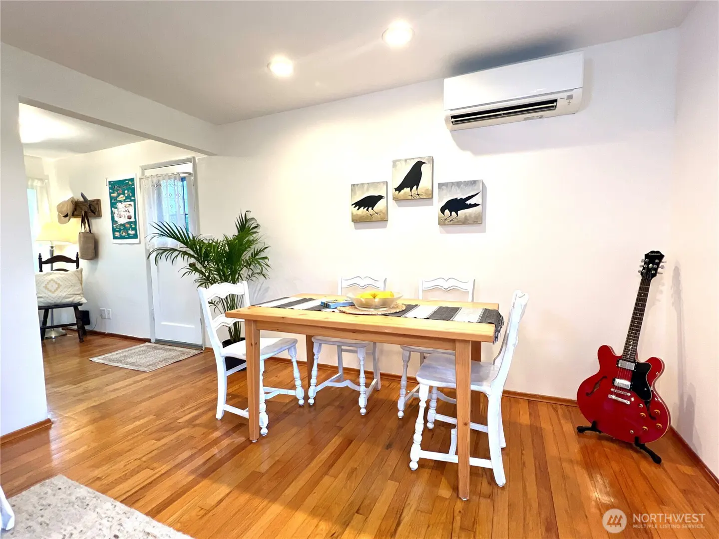 Bright dining room with wood floors, a table with white chairs, and crow artwork on the wall. A red guitar leans against the wall.