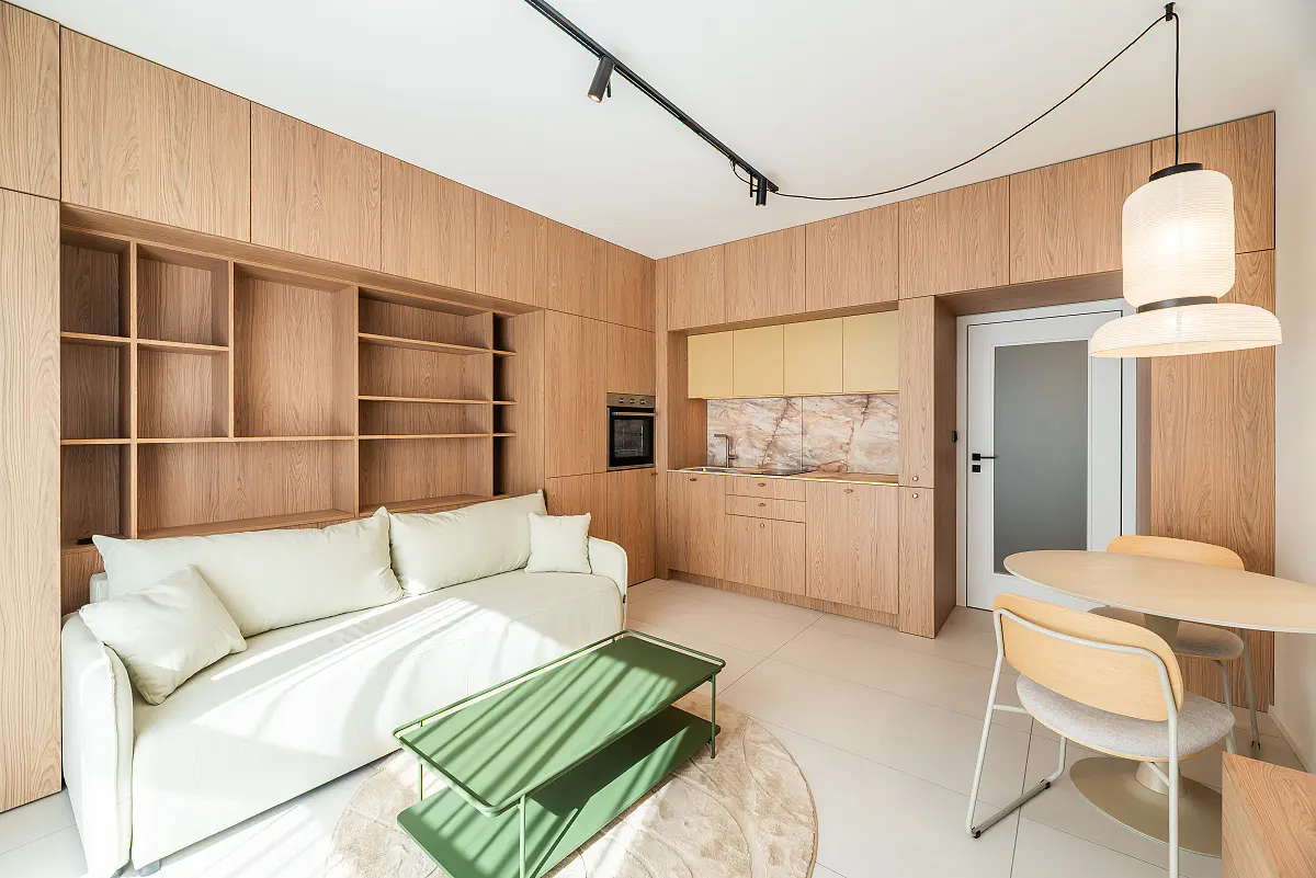 Bright studio apartment with light wood built-ins, white sofa, green coffee table, and round dining table under a modern pendant light.