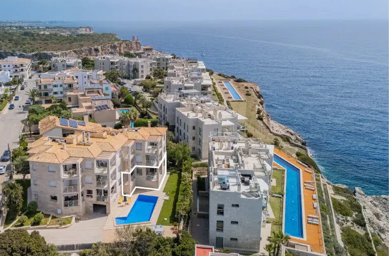 Aerial view of coastal apartments with pools. White buildings with orange roofs line the cliffside overlooking the blue ocean.