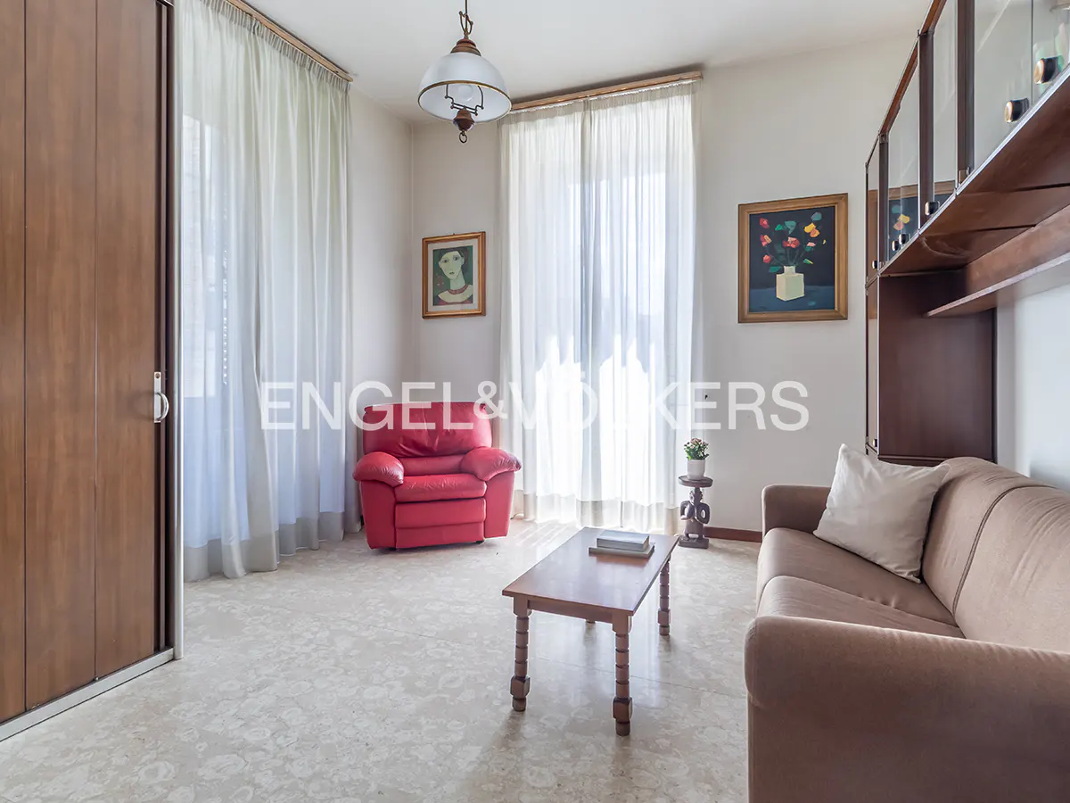 A living room with a brown sofa, red armchair, wood table, and a cabinet. Light streams through sheer white curtains.