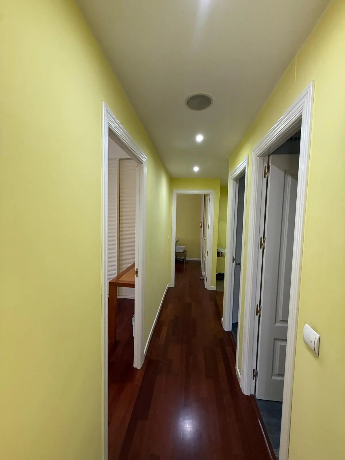 Hallway with yellow walls, dark wood floors, and white trim. Several doorways lead to other rooms. Recessed lighting in the ceiling.
