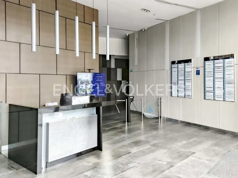 Modern office lobby with a black reception desk, turnstiles, and directory boards on a gray wall. Cylindrical lights hang from the ceiling.