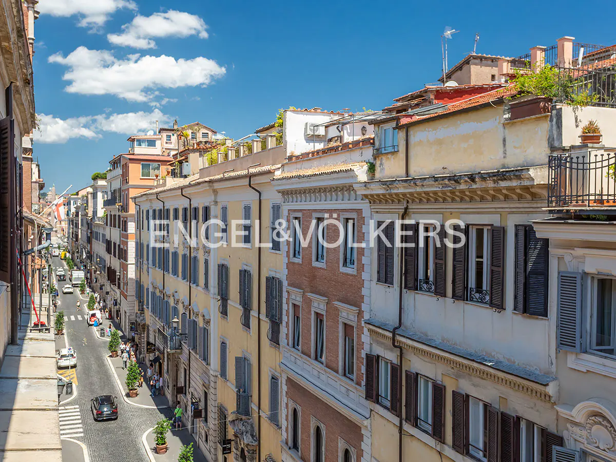 Elevated view of a narrow, cobblestone street in Rome lined with historic, multi-story buildings under a bright blue sky.