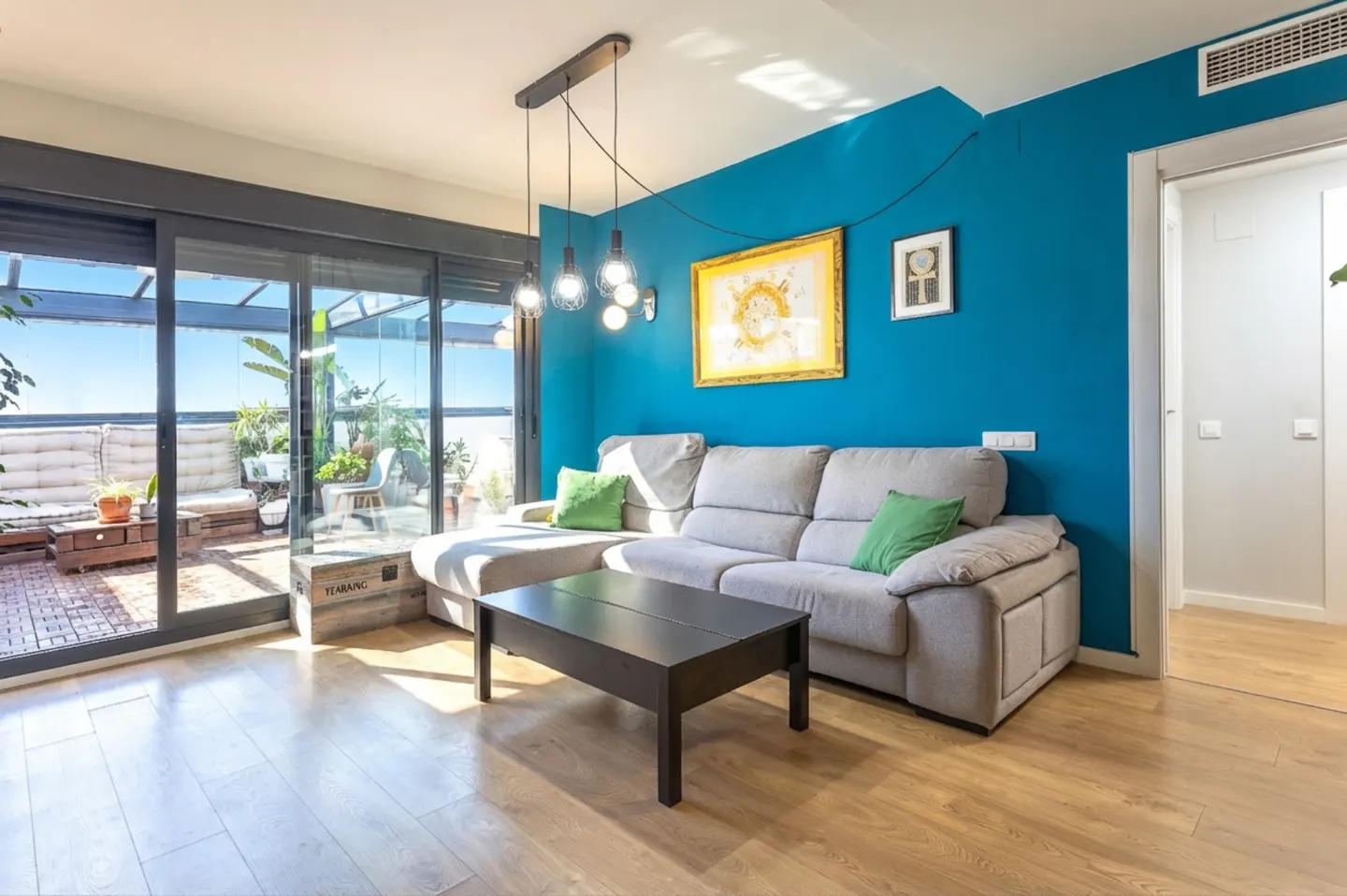 Bright living room with blue accent wall, gray sofa with green pillows, and a black coffee table. Sliding glass doors lead to a sunny patio.