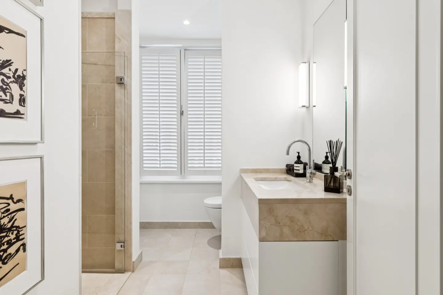 Bright bathroom with white walls, marble countertop, and shuttered window. Artwork hangs on the left wall.