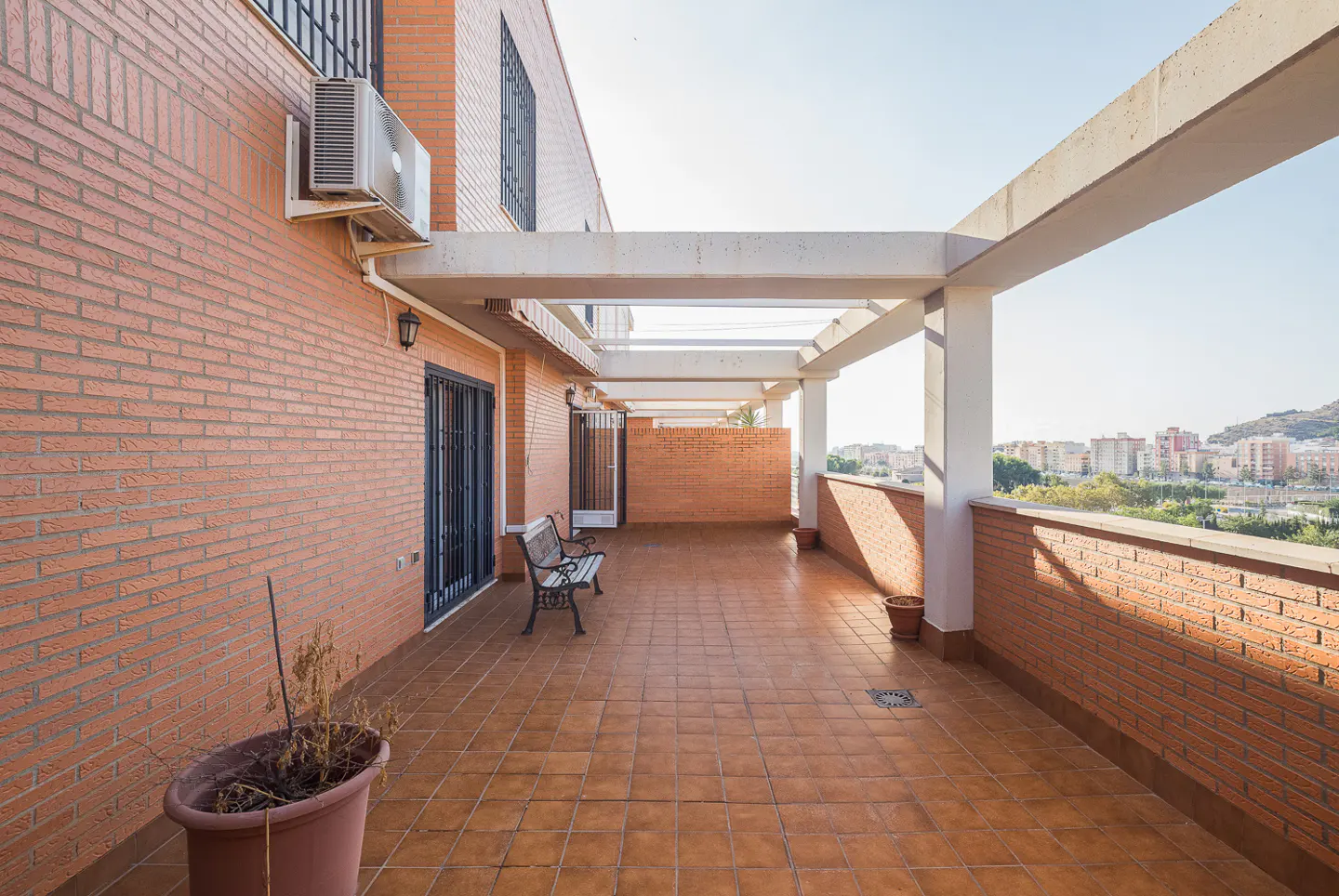 Wide balcony with brick walls, brown tile floor, and white pergola. A bench sits near a black metal door. City view in the background.