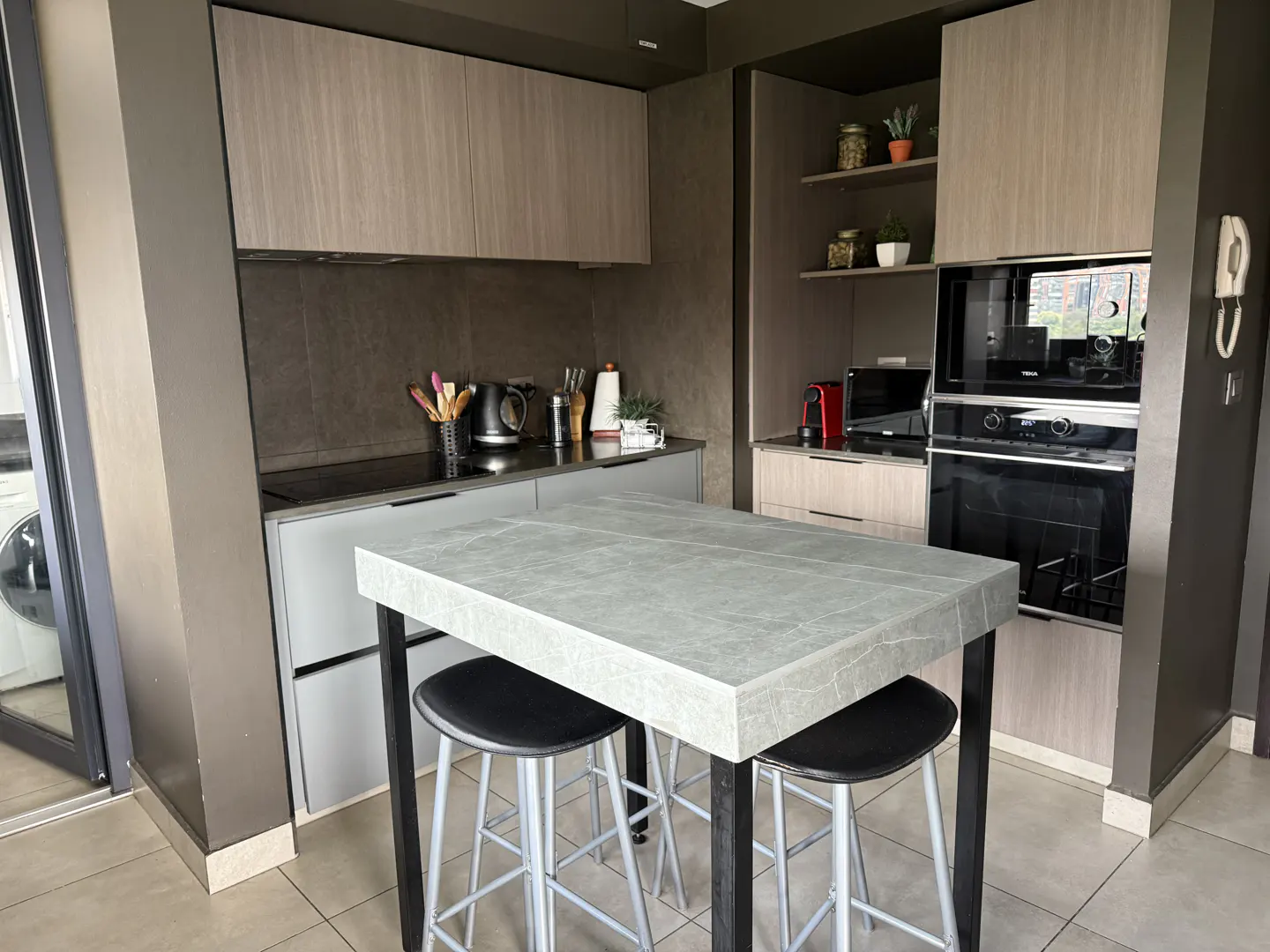 Modern kitchen with light wood cabinets, grey stone island, two black stools, and built-in stainless steel appliances.