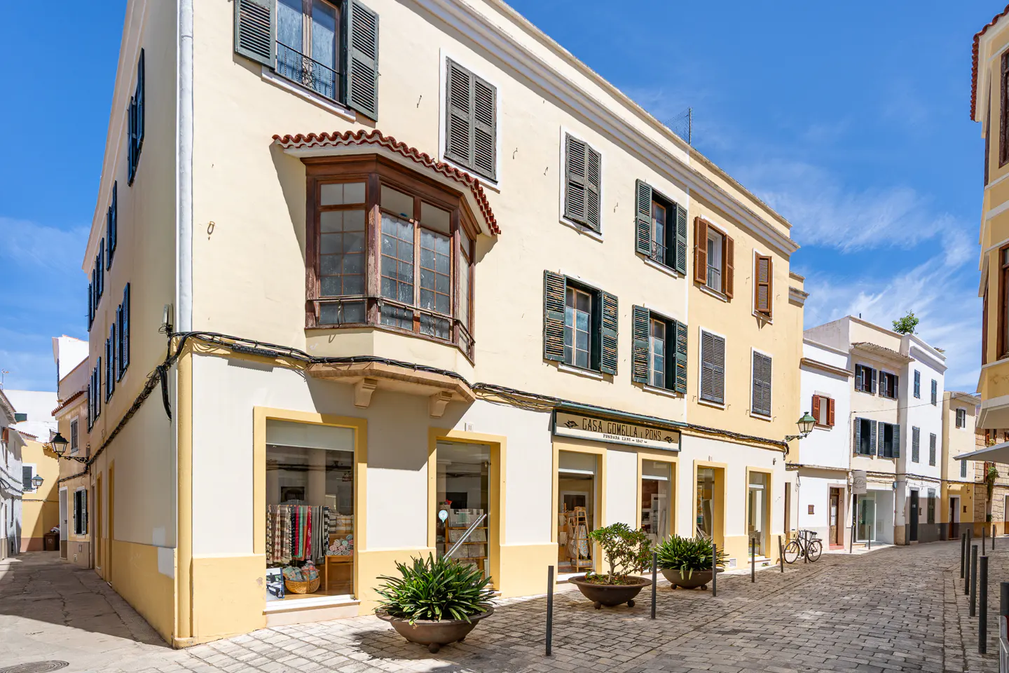 Street view of yellow buildings with shops on the ground floor and shuttered windows under a blue sky.