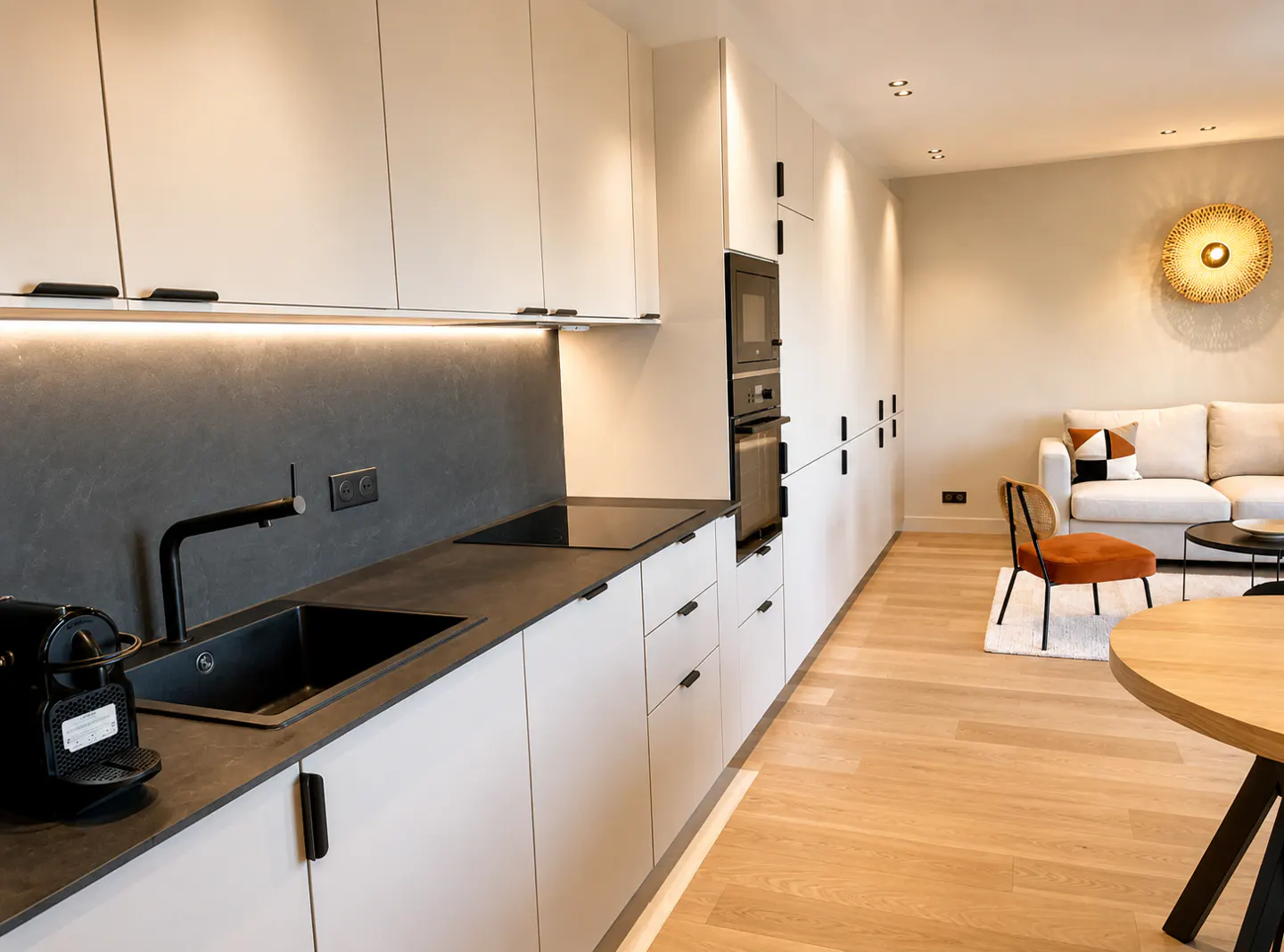 Modern kitchen with white cabinets, black countertops, and stainless steel appliances. Living room with white sofa and round wooden table in background.
