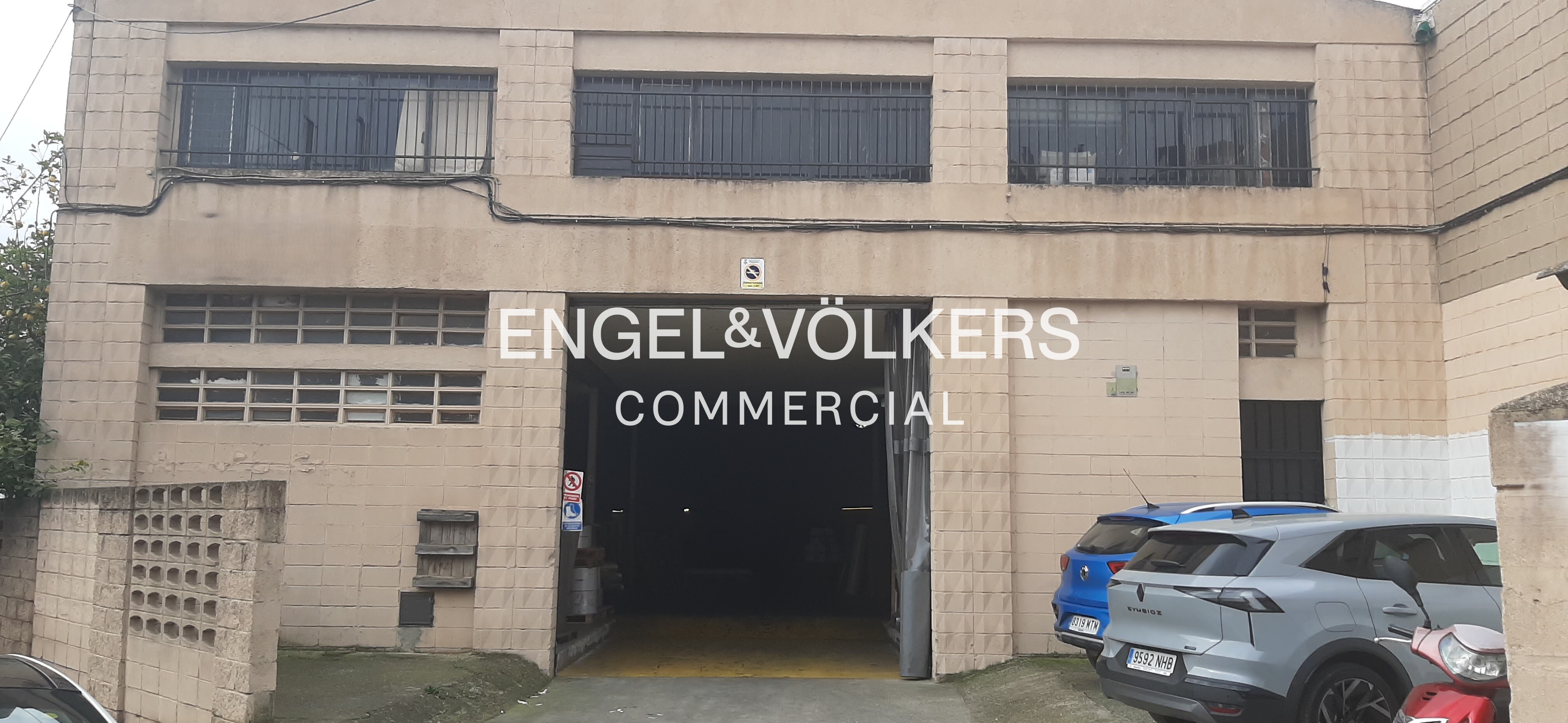 Exterior of a beige industrial warehouse with a large open entrance, featuring parked cars and a scooter in the foreground. [AI-generated text]