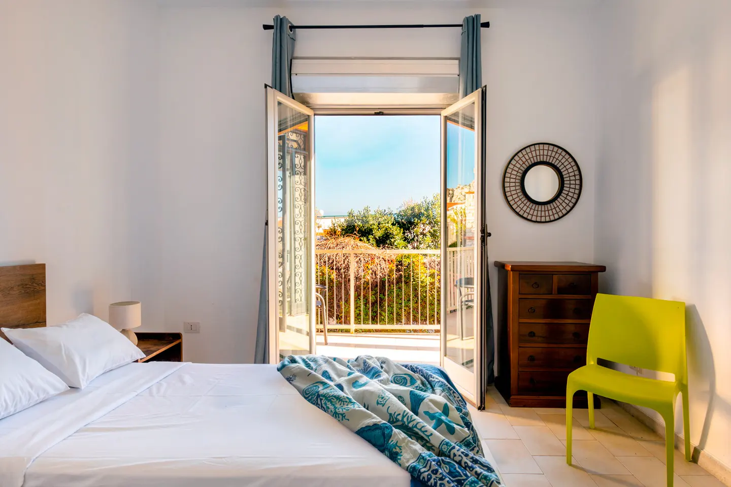 Bedroom with white walls, bed with blue blanket, open balcony doors, wooden dresser, and a green chair.