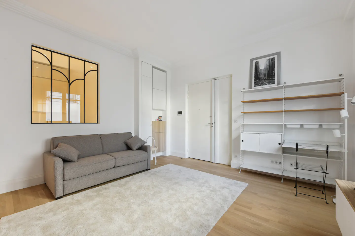 Bright living room with a gray sofa, white rug, and a black-framed window. White shelving unit and a framed print on the wall.