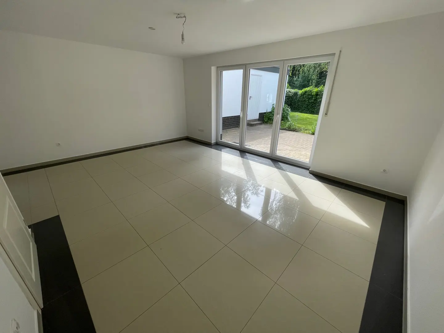 Empty room with white walls, beige tile floor, and black trim. Sliding glass doors lead to a patio and green lawn.