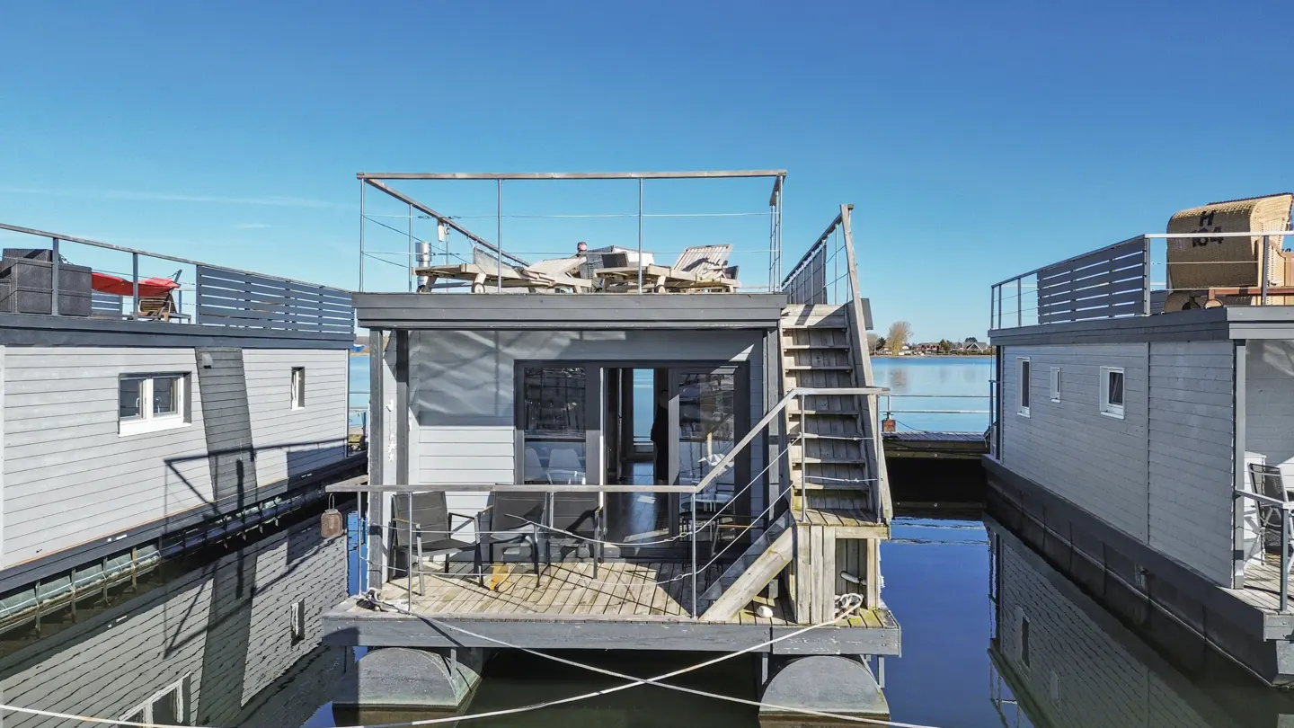 A gray houseboat with a rooftop deck sits on calm water under a blue sky. Stairs lead to the deck with chairs and a table. Other houseboats are nearby.