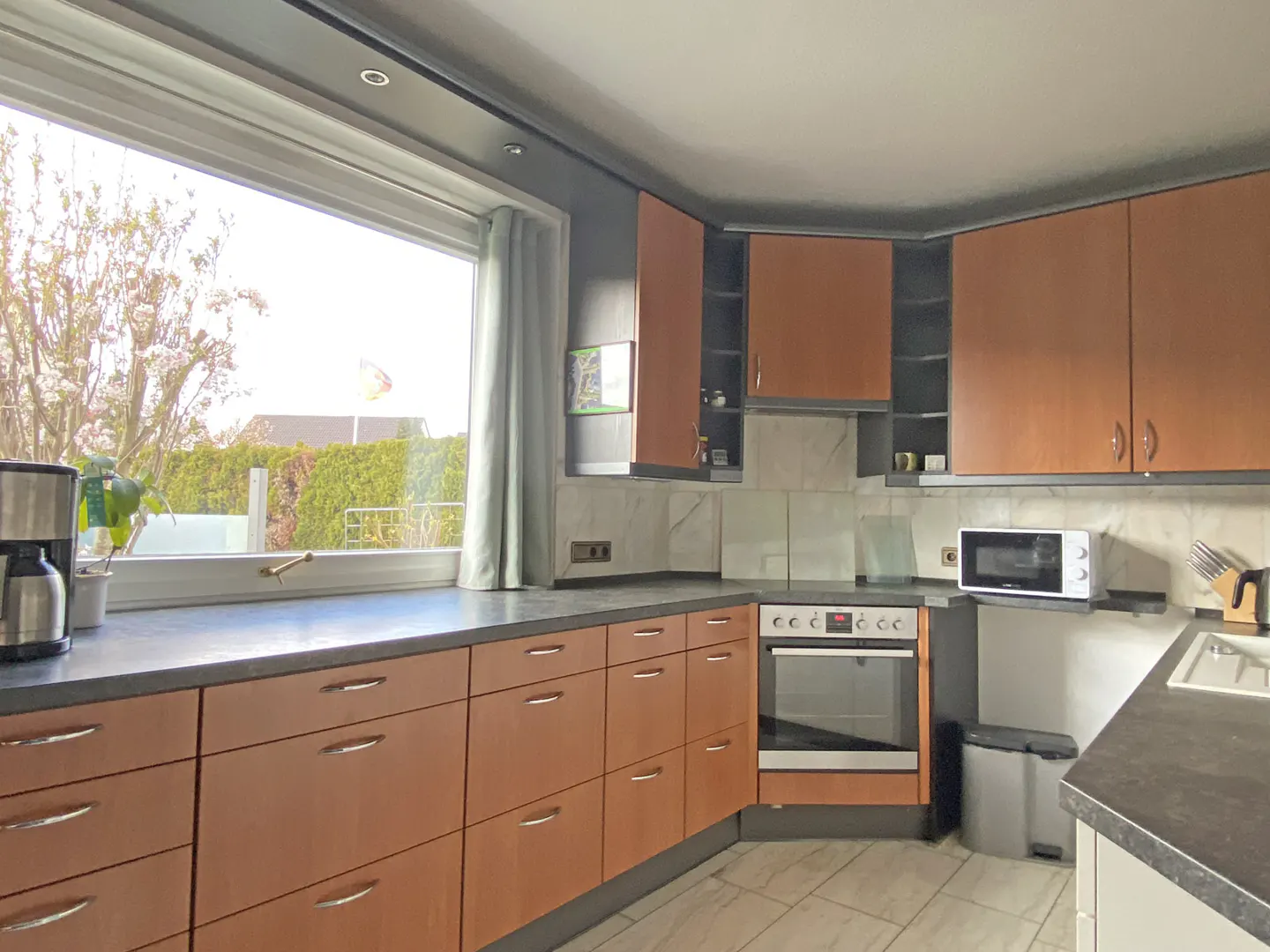 A kitchen with wood cabinets, gray countertops, and a large window. Appliances include an oven, microwave, and coffee maker. Outside is a green hedge.