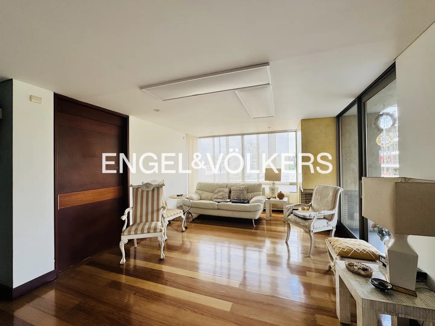 Bright living room with hardwood floors, a white sofa, and two striped chairs. Large windows let in natural light. Engel & Völkers logo visible.