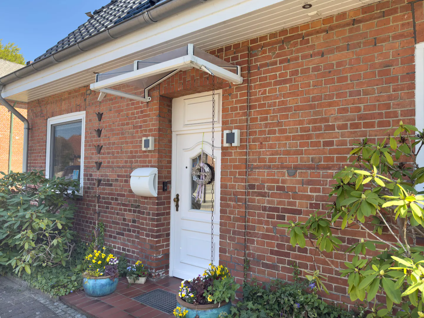 Red brick house with a white door, awning, and mailbox. Flower pots sit on the porch.