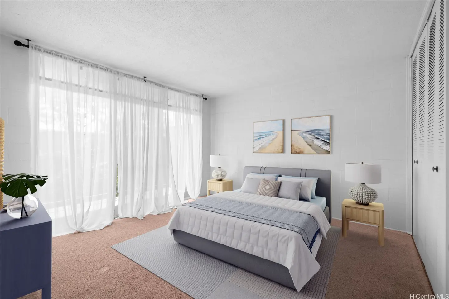 Bedroom with a gray bed, white bedding, and sheer white curtains. Beach-themed art hangs on the white wall. Nightstands with lamps flank the bed.