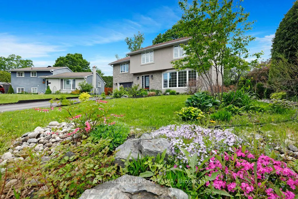Two-story suburban home with stone and stucco exterior, lush green lawn, and a vibrant flower garden in the foreground.