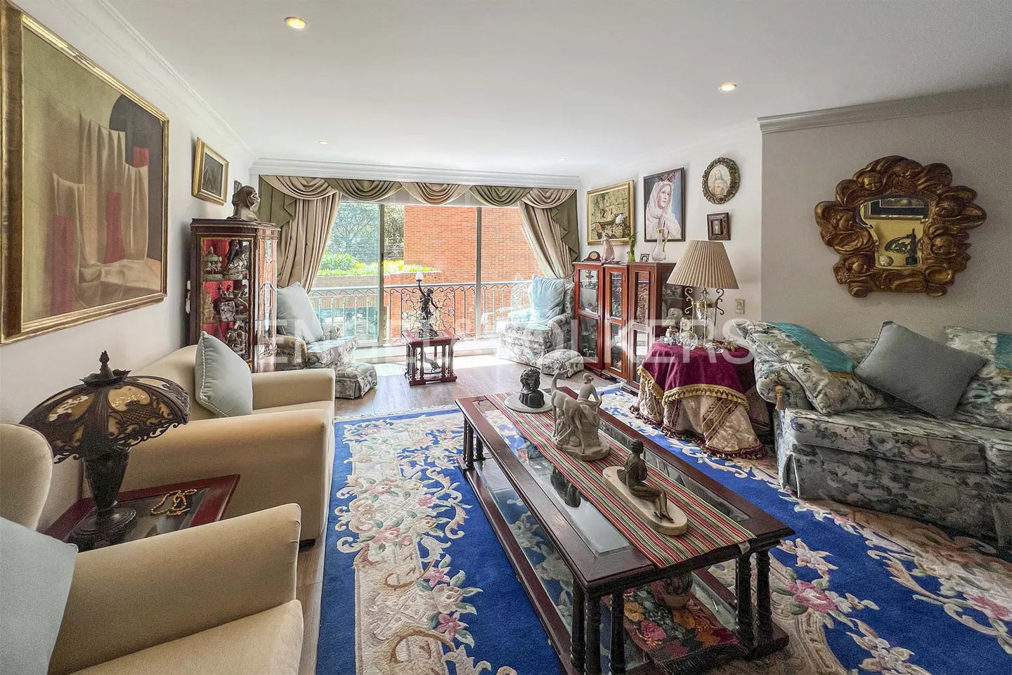 A living room with beige sofas, a blue floral rug, and a glass coffee table. Art and ornate decor fill the space. Balcony access is visible.