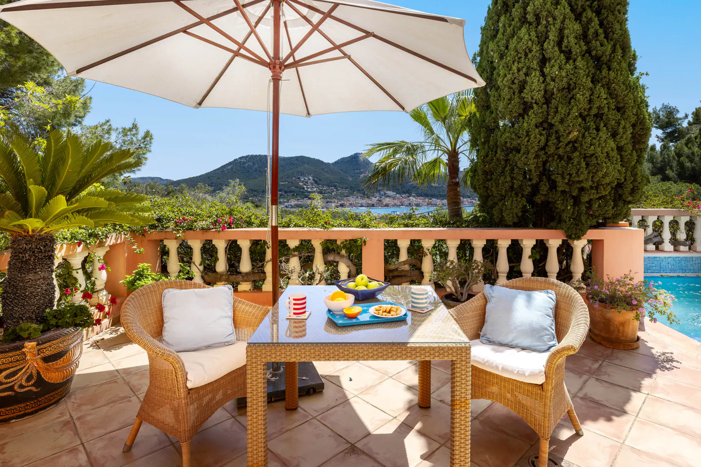 Outdoor patio with wicker chairs, glass table set with fruit, and a white umbrella. A pool and mountain view are in the background.