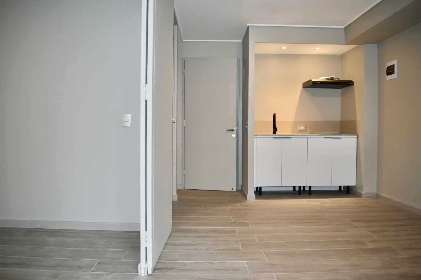 A modern studio apartment with gray walls, light wood-look tile flooring, and a white kitchenette with a black range hood.